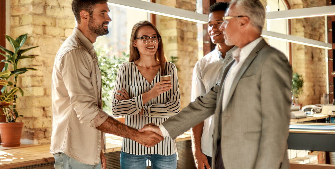 Two business people shaking hands and smiling while standing with coworkers in the creative office.